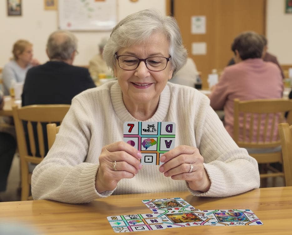A senior woman wearing glasses and a cream sweater smiling while holding a CognaCard at a wooden table in a community setting, playing mind stimulating games with other seniors in the background engaging in similar activities